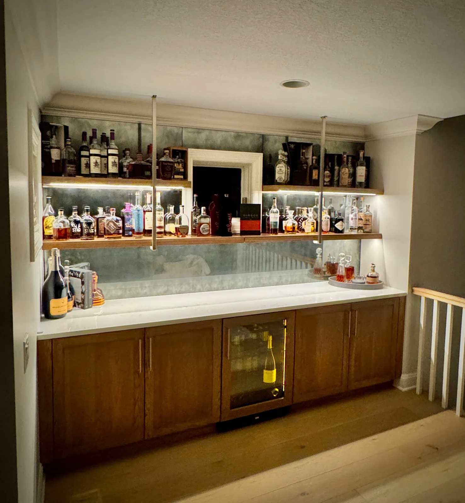 Home bar with a full-height antique mirror backsplash behind two lit floating shelves of bottles.